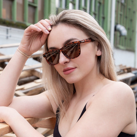 Žena s dugom plavom kosom nosi uniseks naočale SPICY CLASSIC BROWN YELLOW TORTOISE s UV400 zaštitom, sjedi vani uz drvenu površinu, sa zelenim okvirima prozora i drvenim daskama u pozadini.