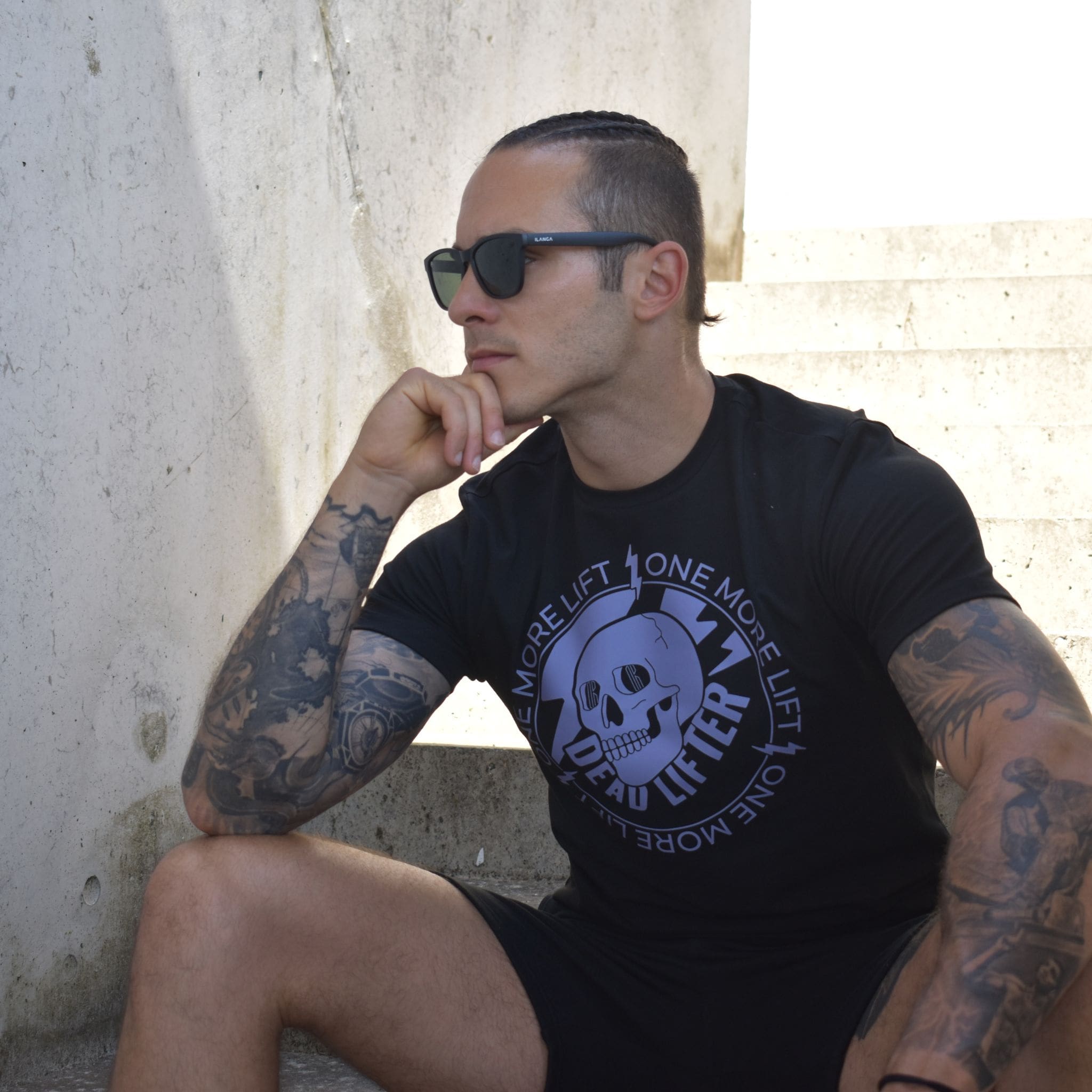 Tetovirana osoba koja nosi sunčane naočale SPICY CLASSIC SMOKE MATTE BLACK s UV400 zaštitom i crnu majicu One More Lift, Deadlifter sjedi na betonskim stepenicama na suncu, naslanjajući bradu na ruku i gledajući u stranu.